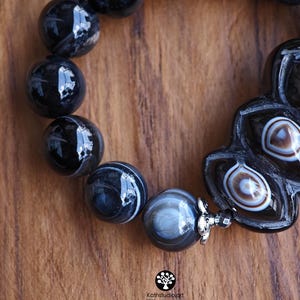 May include: A bracelet featuring polished black and grey banded agate beads, including a larger, uniquely shaped focal bead with concentric white and brown eye-like patterns, accented by a small silver-toned metal flower-shaped spacer bead.