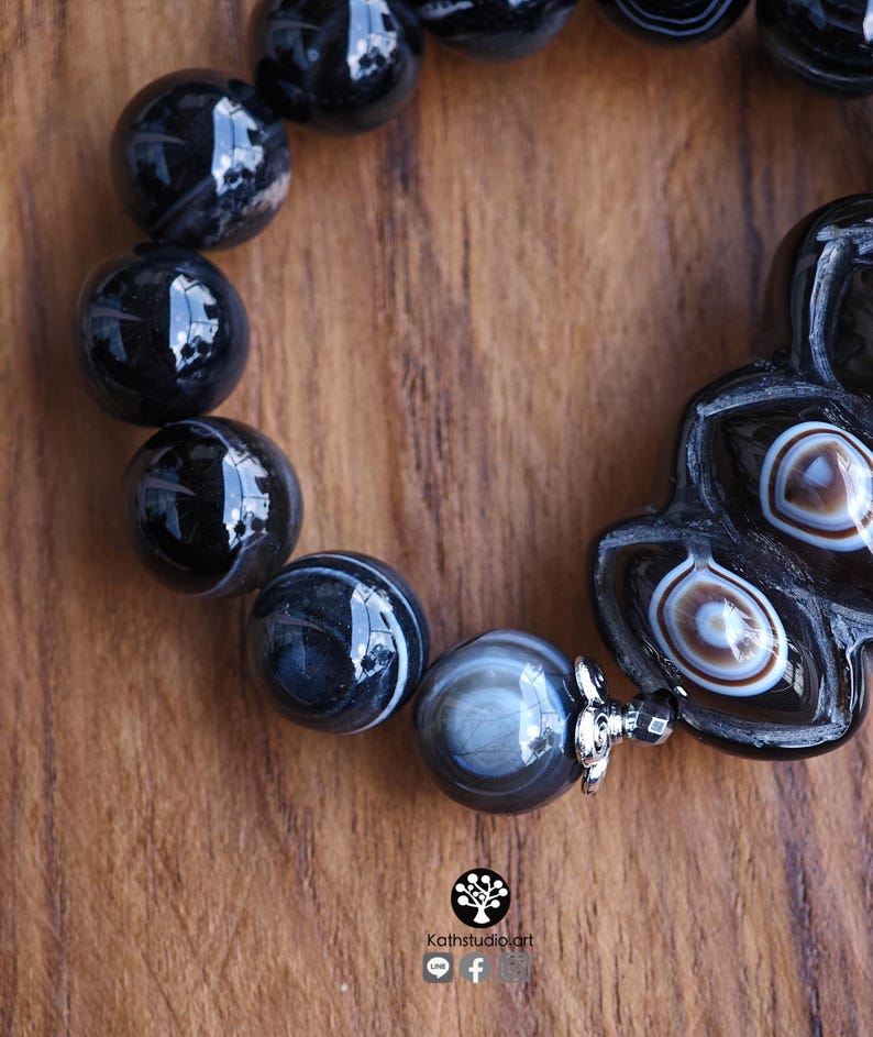May include: A bracelet featuring polished black and grey banded agate beads, including a larger, uniquely shaped focal bead with concentric white and brown eye-like patterns, accented by a small silver-toned metal flower-shaped spacer bead.