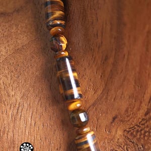 Puede incluir: Un collar con una hilera de cuentas de ojo de tigre, alternando formas cil&iacute;ndricas y esf&eacute;ricas facetadas en tonos marr&oacute;n dorado y marr&oacute;n oscuro. Las cuentas siguen un patr&oacute;n repetitivo. Se observa un logo con el texto Kathstudio.art.