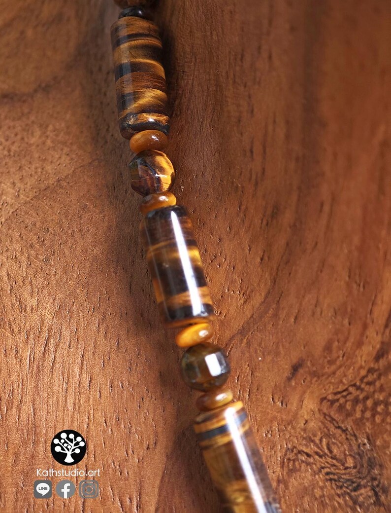 Puede incluir: Un collar con una hilera de cuentas de ojo de tigre, alternando formas cil&iacute;ndricas y esf&eacute;ricas facetadas en tonos marr&oacute;n dorado y marr&oacute;n oscuro. Las cuentas siguen un patr&oacute;n repetitivo. Se observa un logo con el texto Kathstudio.art.