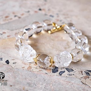 May include: A clear beaded bracelet featuring round transparent beads and two carved floral-shaped beads. The bracelet is accented with small gold-toned metal spacers and a gold-toned toggle clasp. The text Kathstudio.art is visible in the corner.