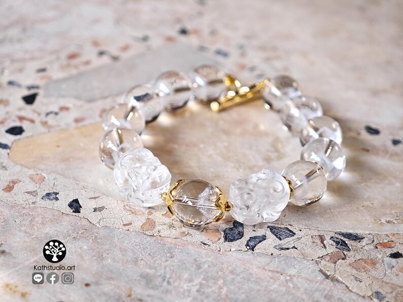 May include: A clear beaded bracelet featuring round transparent beads and two carved floral-shaped beads. The bracelet is accented with small gold-toned metal spacers and a gold-toned toggle clasp. The text Kathstudio.art is visible in the corner.
