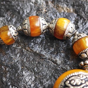 Puede incluir: Una pulsera que presenta cuentas redondas de color &aacute;mbar veteado que se alternan con espaciadores de metal plateado texturizado y ornamentado. Las cuentas muestran un tono c&aacute;lido, naranja y amarillo transl&uacute;cido, mientras que los componentes met&aacute;licos tienen patrones en relieve.
