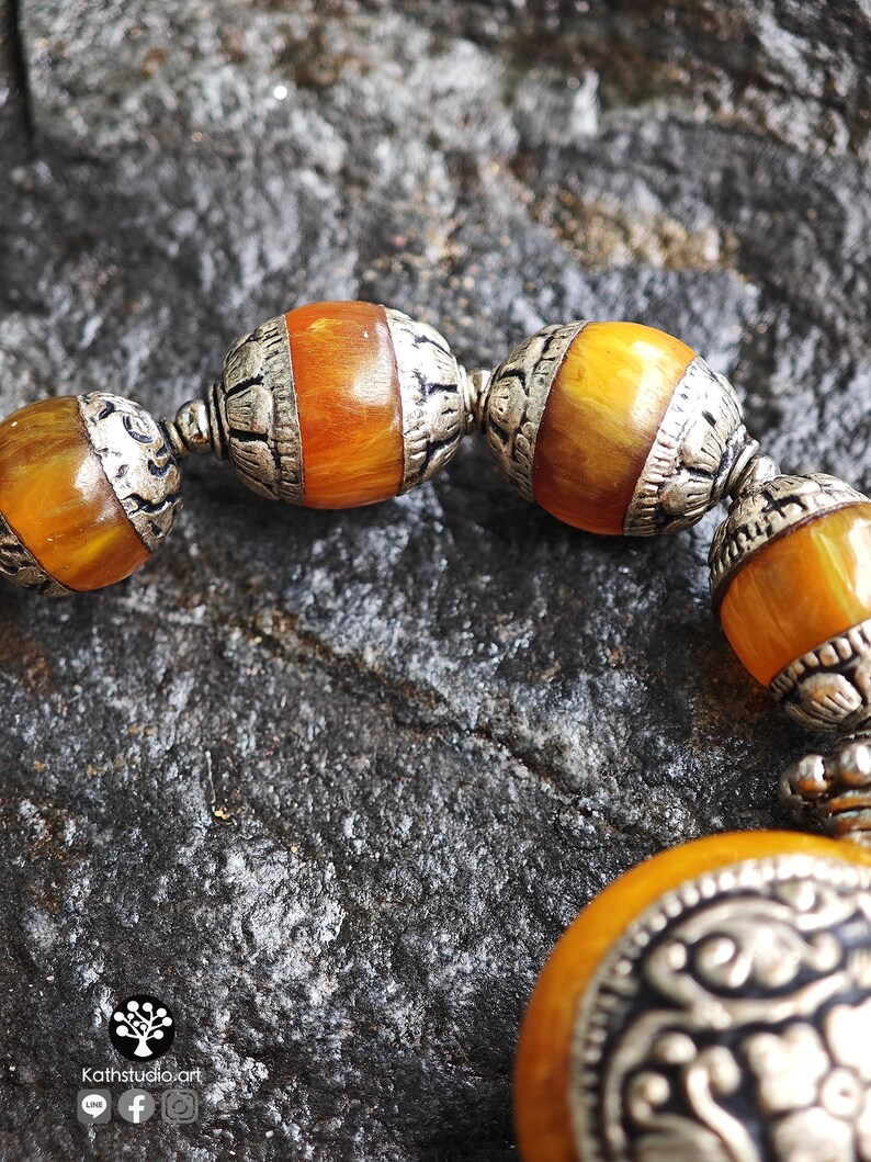 Puede incluir: Una pulsera que presenta cuentas redondas de color &aacute;mbar veteado que se alternan con espaciadores de metal plateado texturizado y ornamentado. Las cuentas muestran un tono c&aacute;lido, naranja y amarillo transl&uacute;cido, mientras que los componentes met&aacute;licos tienen patrones en relieve.