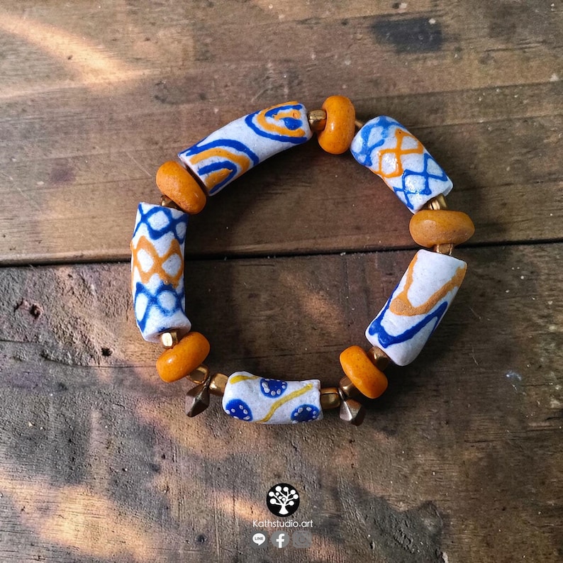 May include: A beaded bracelet featuring cylindrical white beads with blue and orange geometric patterns, interspersed with smaller round mustard-yellow beads and metallic spacers. The design includes a logo and text reading Kathstudio.art.