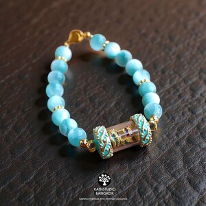K&ouml;nnte beinhalten: Ein Armband mit runden, hellblauen Perlen und kleinen goldfarbenen Zwischenelementen. Das Mittelst&uuml;ck ist ein klarer, zylindrischer Tubus mit einer kleinen goldfarbenen Figur, flankiert von zwei verzierten, t&uuml;rkisfarbenen und goldfarbenen Metallkappen.