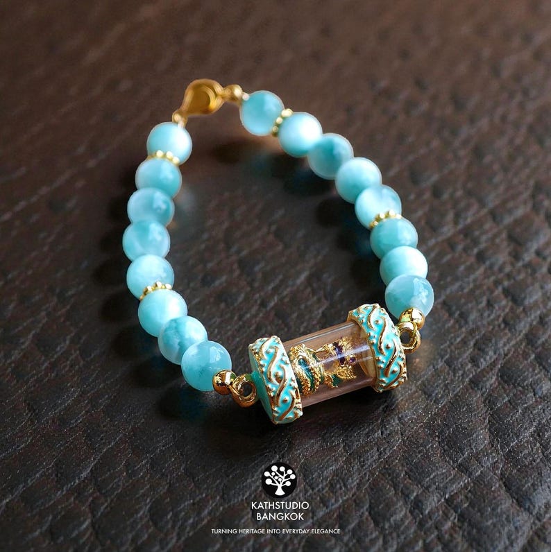 K&ouml;nnte beinhalten: Ein Armband mit runden, hellblauen Perlen und kleinen goldfarbenen Zwischenelementen. Das Mittelst&uuml;ck ist ein klarer, zylindrischer Tubus mit einer kleinen goldfarbenen Figur, flankiert von zwei verzierten, t&uuml;rkisfarbenen und goldfarbenen Metallkappen.
