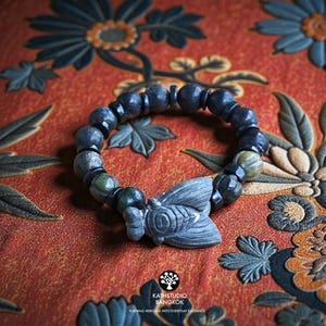 Puede incluir: Una pulsera de cuentas con piedras de color gris oscuro y verde dispuestas en un patr&oacute;n circular. Un dije de piedra gris tallada con forma de cigarra se encuentra en el centro de la pulsera. Incluye peque&ntilde;as cuentas espaciadoras negras.