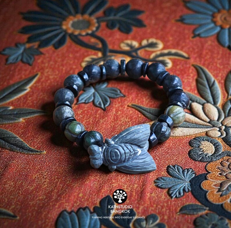 Puede incluir: Una pulsera de cuentas con piedras de color gris oscuro y verde dispuestas en un patr&oacute;n circular. Un dije de piedra gris tallada con forma de cigarra se encuentra en el centro de la pulsera. Incluye peque&ntilde;as cuentas espaciadoras negras.