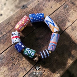 May include: A bracelet composed of cylindrical beads in various colors including red, blue, white, green, and brown, each featuring distinct hand-painted patterns such as dots, stripes, and swirls, separated by small metallic spacers and a bell charm.