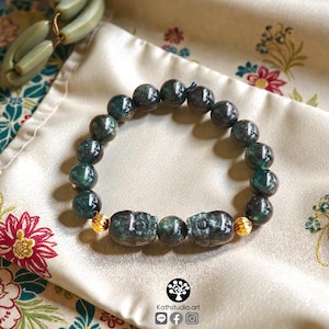 May include: A beaded bracelet featuring dark green, mottled stone beads arranged in a circular pattern. The design includes two carved stone beads shaped like beetles, separated by a single round bead, and accented with two small, textured gold-tone metal beads.