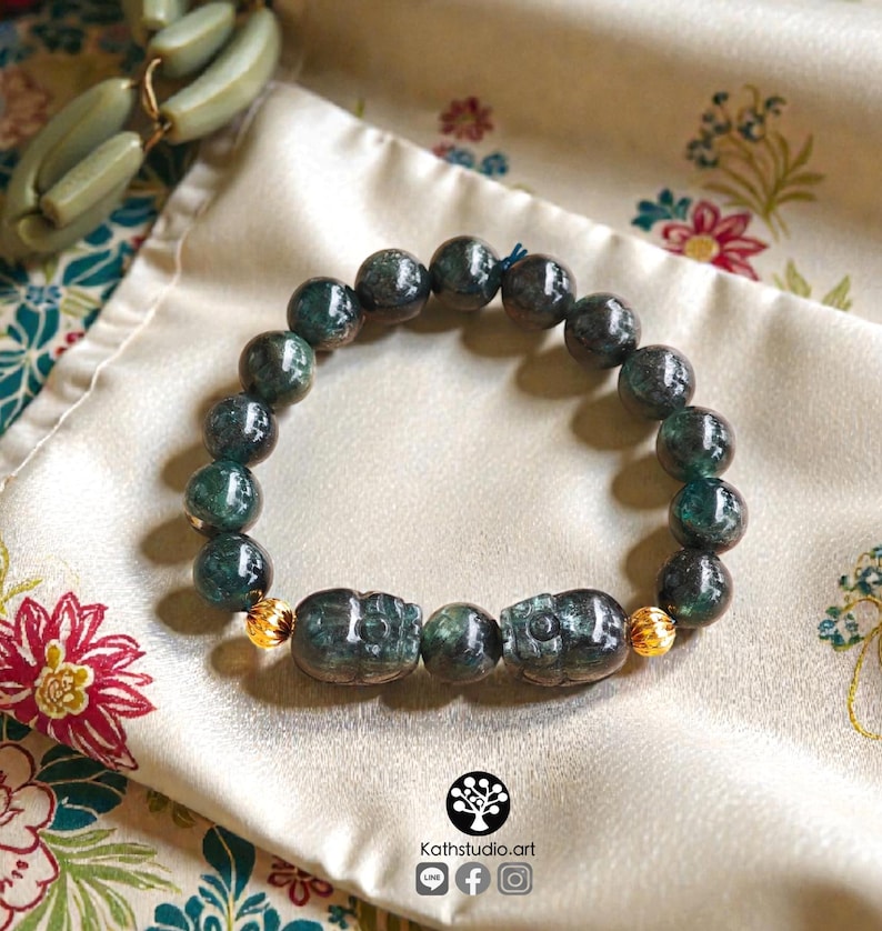 May include: A beaded bracelet featuring dark green, mottled stone beads arranged in a circular pattern. The design includes two carved stone beads shaped like beetles, separated by a single round bead, and accented with two small, textured gold-tone metal beads.