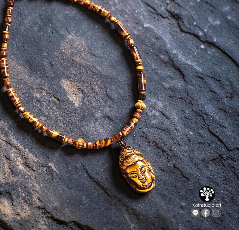 Puede incluir: Un collar con una serie de cuentas cil&iacute;ndricas y esf&eacute;ricas de ojo de tigre en tonos marr&oacute;n y dorado. El collar incluye un colgante de ojo de tigre tallado que representa un rostro sereno con los ojos cerrados y un tocado ornamentado.