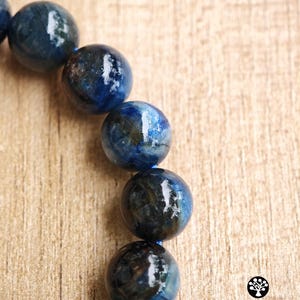 Pu&ograve; includere: Un bracciale composto da perle sferiche lucide di cianite blu con inclusioni scure e marmorizzate, infilate su un cordino blu. Una singola perla metallica testurizzata color oro &egrave; posizionata verso il basso. Il testo Kathstudio.art &egrave; visibile.
