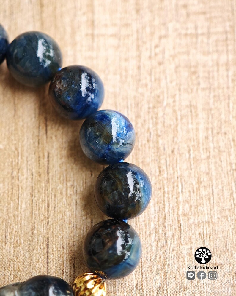 Pu&ograve; includere: Un bracciale composto da perle sferiche lucide di cianite blu con inclusioni scure e marmorizzate, infilate su un cordino blu. Una singola perla metallica testurizzata color oro &egrave; posizionata verso il basso. Il testo Kathstudio.art &egrave; visibile.