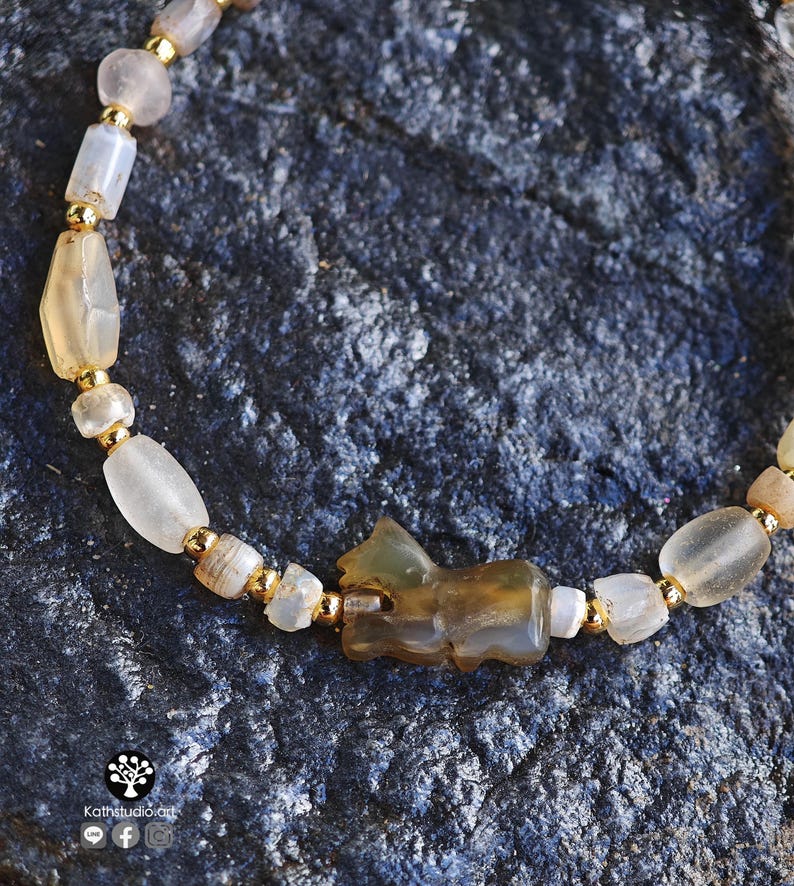 May include: A beaded bracelet featuring translucent, irregularly shaped light-colored stone beads interspersed with small, round, metallic gold-tone spacer beads. A central carved stone animal figure is positioned among the beads.