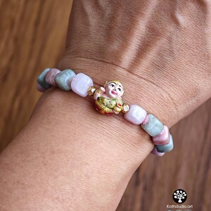 May include: A beaded bracelet featuring alternating square-shaped light pink and sage green stones. The centerpiece is a small, detailed gold-toned figurine of a smiling child wearing a green and red garment, positioned between the stone beads.