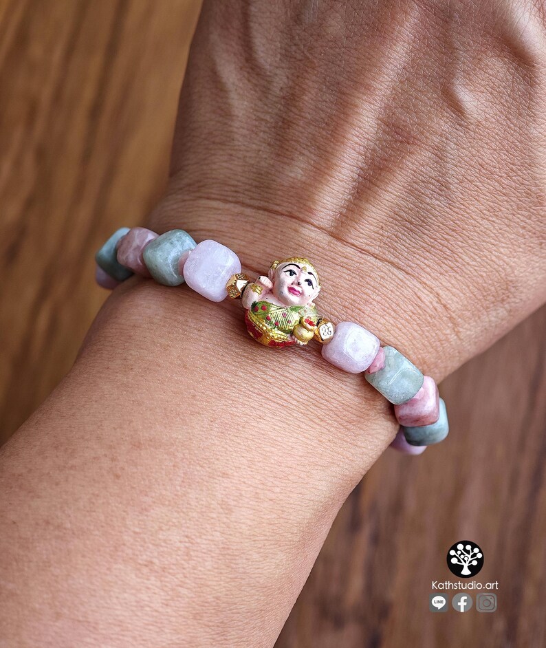May include: A beaded bracelet featuring alternating square-shaped light pink and sage green stones. The centerpiece is a small, detailed gold-toned figurine of a smiling child wearing a green and red garment, positioned between the stone beads.