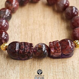 Peut inclure: Un bracelet compos&eacute; de perles sph&eacute;riques polies, rouge fonc&eacute; et translucides, ainsi que de deux perles sculpt&eacute;es en forme d'animal. Le bracelet comporte de petites entretoises m&eacute;talliques dor&eacute;es et stri&eacute;es. Le texte Kathstudio.art est imprim&eacute; en bas.