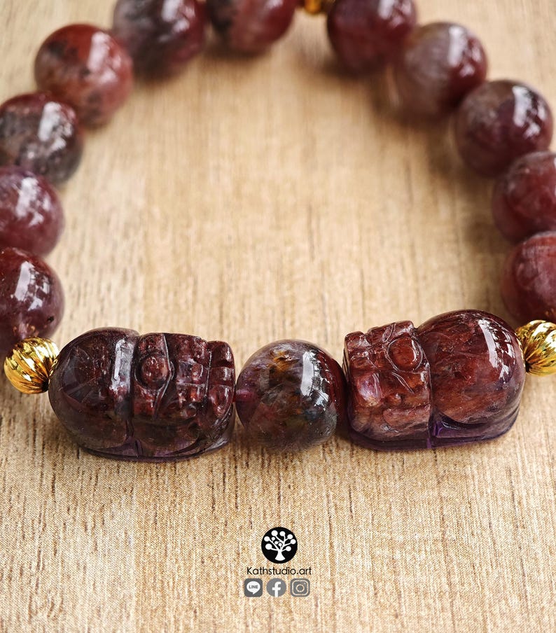 Peut inclure: Un bracelet compos&eacute; de perles sph&eacute;riques polies, rouge fonc&eacute; et translucides, ainsi que de deux perles sculpt&eacute;es en forme d'animal. Le bracelet comporte de petites entretoises m&eacute;talliques dor&eacute;es et stri&eacute;es. Le texte Kathstudio.art est imprim&eacute; en bas.