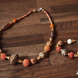 May include: A necklace and bracelet set featuring a variety of beads in shades of brown, orange, and cream. The necklace has a mix of round and oblong beads, with a large, textured stone-like bead. The bracelet mirrors the necklace's design.
