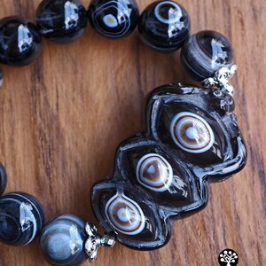 May include: A bracelet featuring polished black agate beads with white and brown concentric eye patterns. The centerpiece is a large, elongated, carved black bead with three distinct eye-like markings, accented by silver-toned metal bead caps.