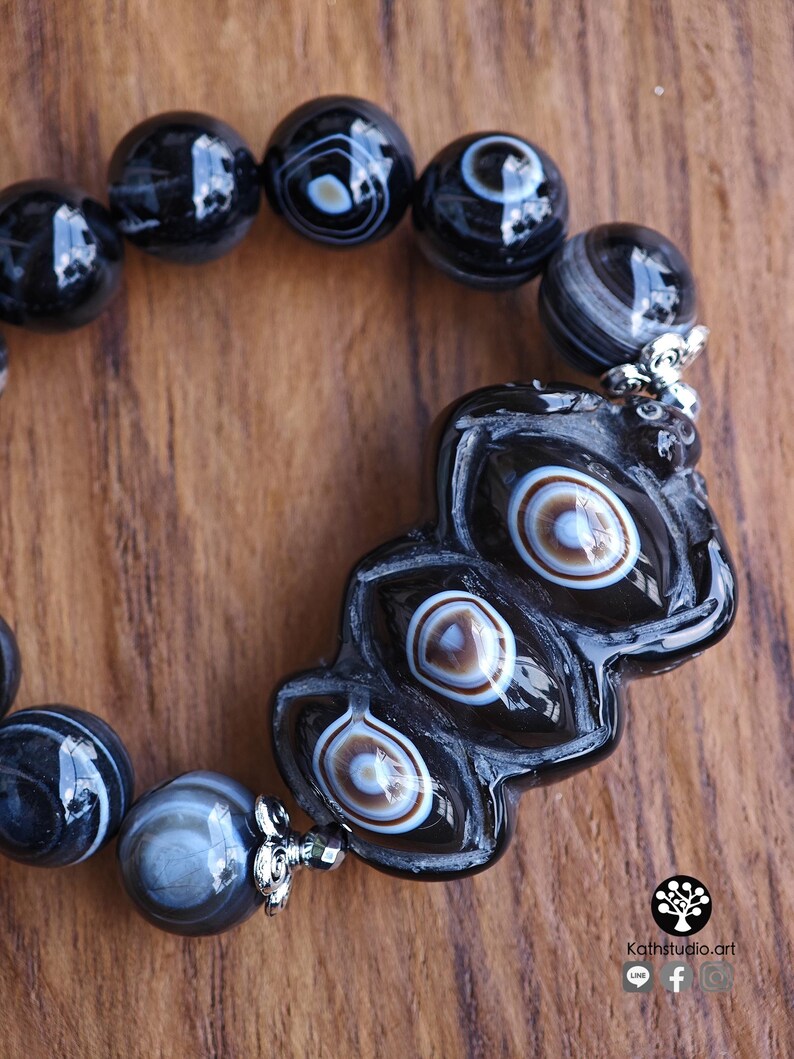 May include: A bracelet featuring polished black agate beads with white and brown concentric eye patterns. The centerpiece is a large, elongated, carved black bead with three distinct eye-like markings, accented by silver-toned metal bead caps.