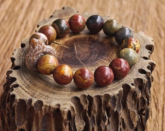 May include: A beaded bracelet featuring round, multi-colored jasper stones in shades of red, brown, green, and yellow, accented by a carved stone dragon charm. The text KATHSTUDIO BANGKOK TURNING HERITAGE INTO EVERYDAY ELEGANCE is printed at the bottom.