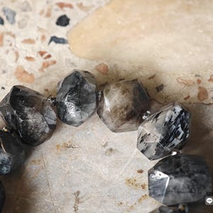May include: A bracelet composed of large, faceted, irregular beads in shades of charcoal, black, and translucent gray with white inclusions. The beads are strung together with small, metallic, dark-colored spacer beads between each larger stone.