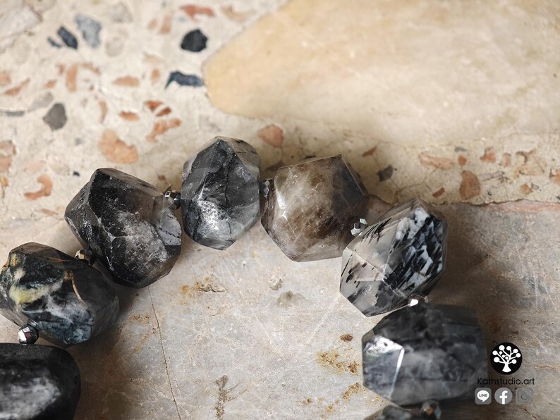 May include: A bracelet composed of large, faceted, irregular beads in shades of charcoal, black, and translucent gray with white inclusions. The beads are strung together with small, metallic, dark-colored spacer beads between each larger stone.