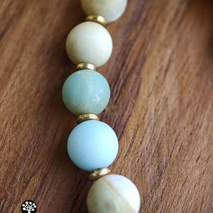 May include: A beaded necklace featuring alternating round matte beads in shades of pale blue, cream, and soft green, separated by small metallic gold-toned spacers. The beads display natural variations in color and texture throughout the strand.