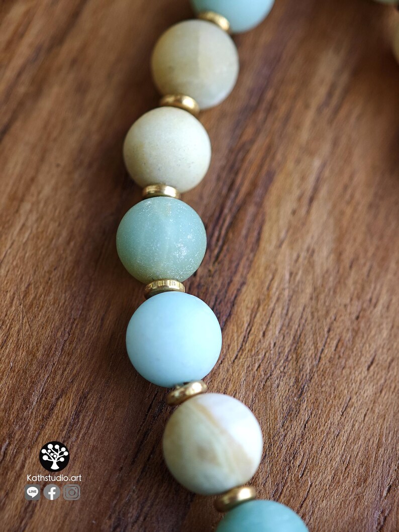 May include: A beaded necklace featuring alternating round matte beads in shades of pale blue, cream, and soft green, separated by small metallic gold-toned spacers. The beads display natural variations in color and texture throughout the strand.