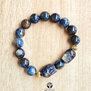 Pu&ograve; includere: Un braccialetto di perline composto da pietre rotonde di cianite blu lucidata con visibili inclusioni scure. Il design include due perline intagliate a forma di teschio e due piccole perline distanziatrici in metallo dorato testurizzato.