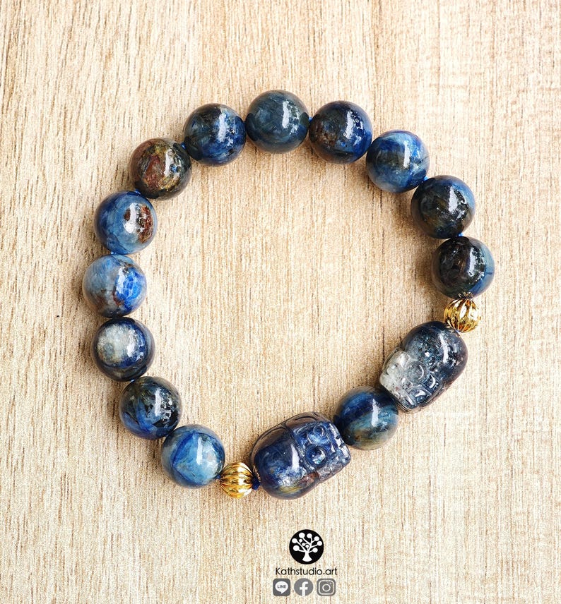 Pu&ograve; includere: Un braccialetto di perline composto da pietre rotonde di cianite blu lucidata con visibili inclusioni scure. Il design include due perline intagliate a forma di teschio e due piccole perline distanziatrici in metallo dorato testurizzato.