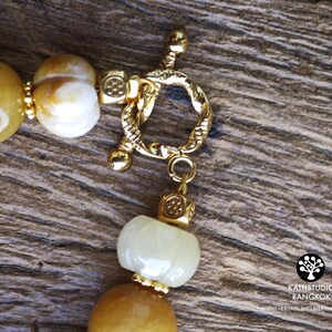 Op de afbeelding: Een ketting met gladde, ronde gele en witte kralen, geaccentueerd met kleine goudkleurige vierkante tussenstukjes en een getextureerde goudkleurige knevelsluiting. Het logo KATHSTUDIO BANGKOK en de tekst TURNING HERITAGE INTO EVERYDAY ELEGANCE zijn zichtbaar.