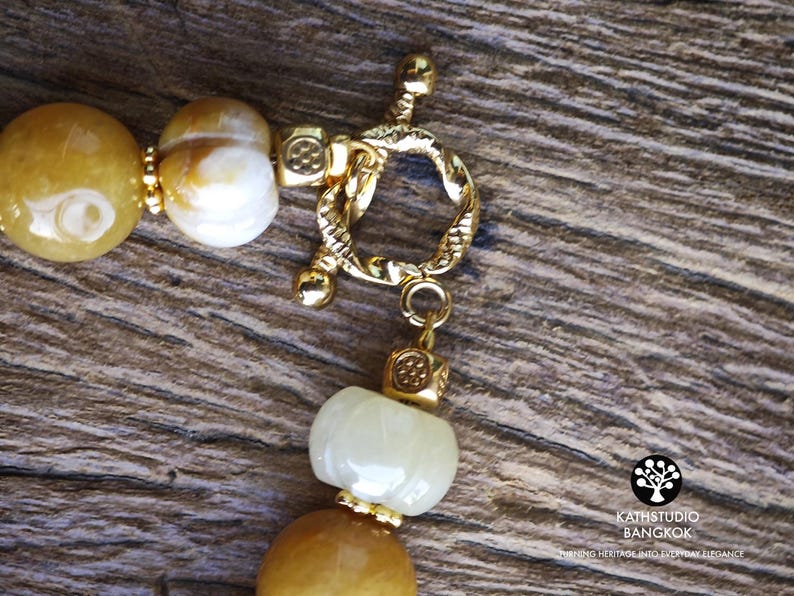 Op de afbeelding: Een ketting met gladde, ronde gele en witte kralen, geaccentueerd met kleine goudkleurige vierkante tussenstukjes en een getextureerde goudkleurige knevelsluiting. Het logo KATHSTUDIO BANGKOK en de tekst TURNING HERITAGE INTO EVERYDAY ELEGANCE zijn zichtbaar.