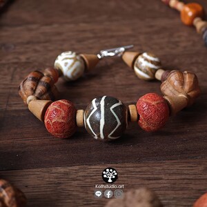 May include: A handcrafted bracelet with a variety of wooden beads in shades of brown, red, and white. The bracelet features a central bead with a unique white pattern and a silver clasp. The beads are strung together with a natural cord.