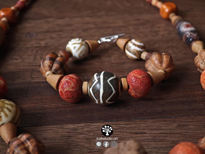 May include: A handcrafted bracelet with a variety of wooden beads in shades of brown, red, and white. The bracelet features a central bead with a unique white pattern and a silver clasp. The beads are strung together with a natural cord.