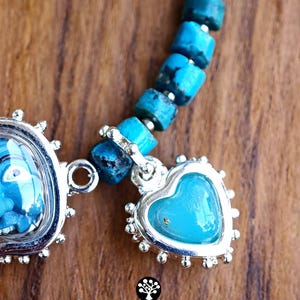Puede incluir: Una pulsera con cuentas cil&iacute;ndricas de turquesa y un dije plateado en forma de coraz&oacute;n con un centro azul liso. El dije est&aacute; enmarcado por un borde festoneado decorativo. El texto KATHSTUDIO BANGKOK est&aacute; impreso debajo de la joya.