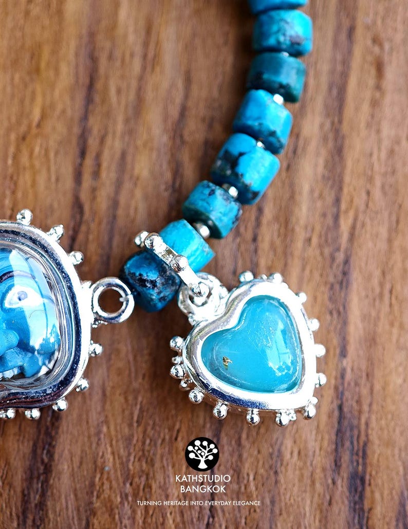 Puede incluir: Una pulsera con cuentas cil&iacute;ndricas de turquesa y un dije plateado en forma de coraz&oacute;n con un centro azul liso. El dije est&aacute; enmarcado por un borde festoneado decorativo. El texto KATHSTUDIO BANGKOK est&aacute; impreso debajo de la joya.
