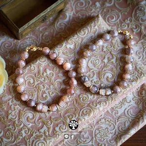 May include: Two beaded bracelets featuring round, polished sunstone beads in shades of peach, tan, and brown. Each bracelet includes small gold-toned metal spacers and a gold-toned magnetic clasp. One bracelet features a carved sunstone skull bead.