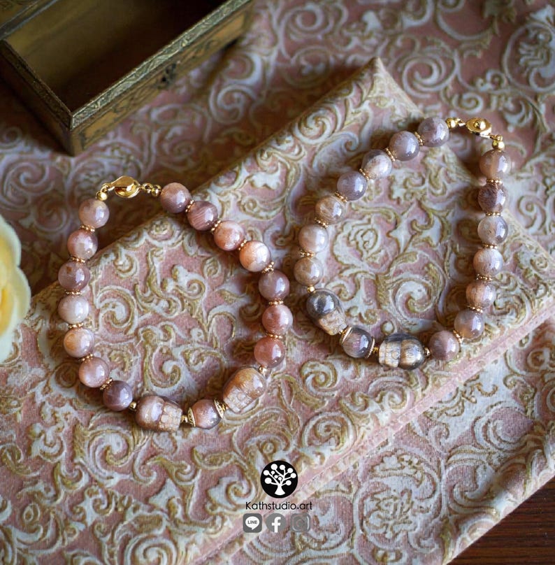 May include: Two beaded bracelets featuring round, polished sunstone beads in shades of peach, tan, and brown. Each bracelet includes small gold-toned metal spacers and a gold-toned magnetic clasp. One bracelet features a carved sunstone skull bead.