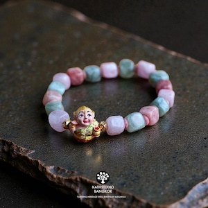 May include: A stretch bracelet featuring alternating smooth, rounded square beads in shades of pale pink and soft green. A central gold-toned charm depicts a small figure with a painted face and green clothing, flanked by two small gold beads.