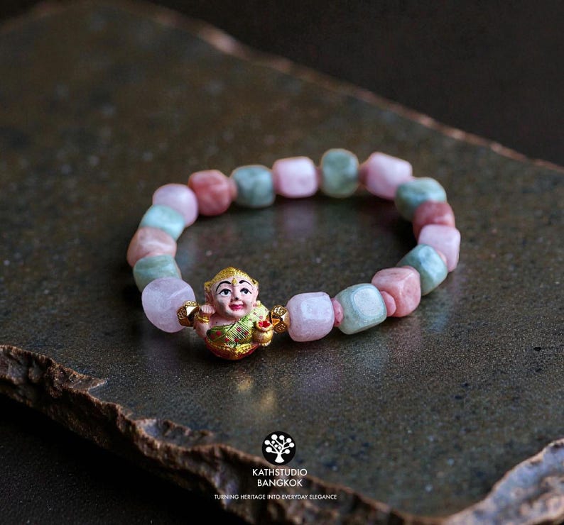 May include: A stretch bracelet featuring alternating smooth, rounded square beads in shades of pale pink and soft green. A central gold-toned charm depicts a small figure with a painted face and green clothing, flanked by two small gold beads.