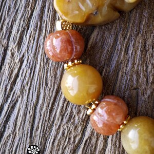 Op de afbeelding: Een armband met gladde gele en oranje stenen kralen, waaronder uitgesneden pompoenvormige kralen en een grotere gele stenen bedel. Kleine goudkleurige metalen tussenstukjes bevinden zich tussen de kralen. Het logo KATHSTUDIO BANGKOK is zichtbaar.