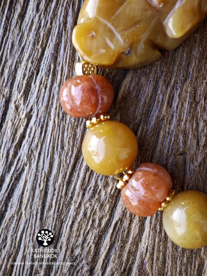 Op de afbeelding: Een armband met gladde gele en oranje stenen kralen, waaronder uitgesneden pompoenvormige kralen en een grotere gele stenen bedel. Kleine goudkleurige metalen tussenstukjes bevinden zich tussen de kralen. Het logo KATHSTUDIO BANGKOK is zichtbaar.