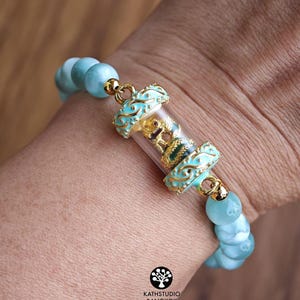 K&ouml;nnte beinhalten: Ein Armband mit hellblauen runden Perlen und einer zentralen, durchsichtigen zylindrischen Kapsel, die eine kleine goldfarbene Figur enth&auml;lt. Die Kapsel ist mit verzierten hellblauen und goldenen Metallelementen versehen. Der Text KATHSTUDIO BANGKOK ist sichtbar.