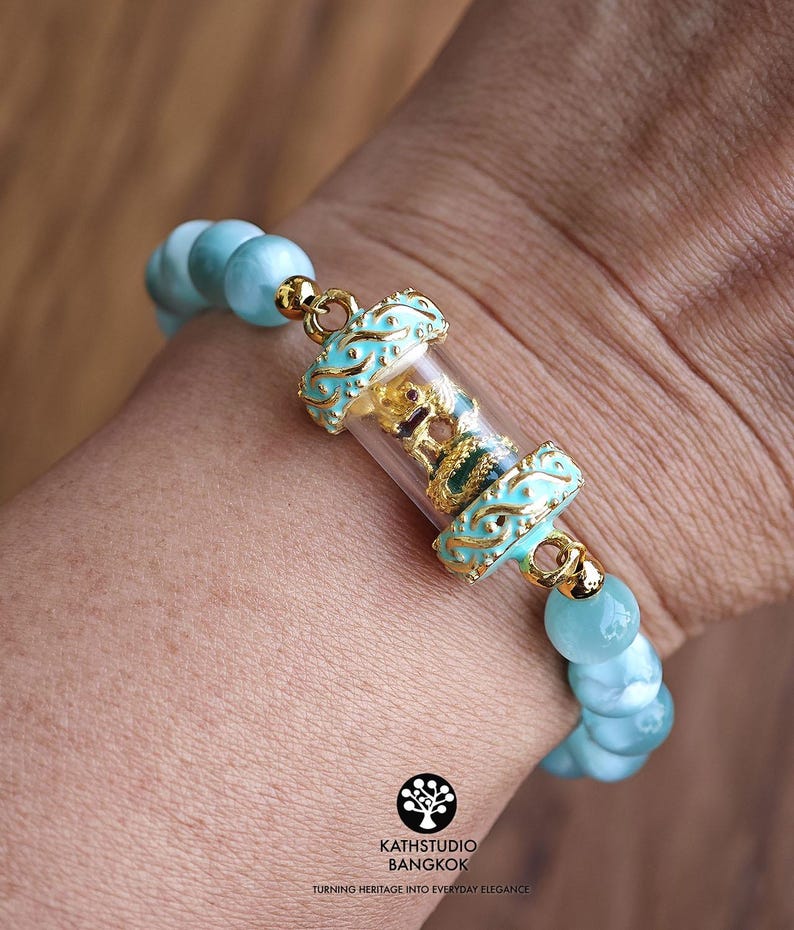 K&ouml;nnte beinhalten: Ein Armband mit hellblauen runden Perlen und einer zentralen, durchsichtigen zylindrischen Kapsel, die eine kleine goldfarbene Figur enth&auml;lt. Die Kapsel ist mit verzierten hellblauen und goldenen Metallelementen versehen. Der Text KATHSTUDIO BANGKOK ist sichtbar.