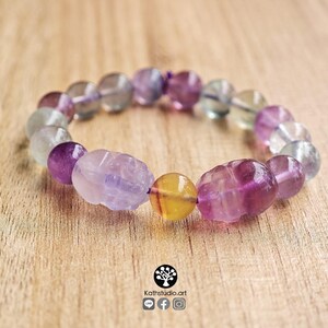 May include: A stretch bracelet composed of round and carved skull-shaped fluorite beads in shades of purple, green, and yellow. The beads are translucent and vary in color intensity, featuring a central yellow bead flanked by two carved purple skulls.