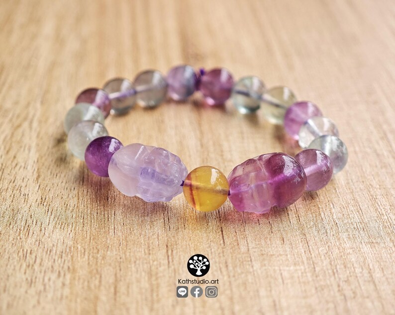 May include: A stretch bracelet composed of round and carved skull-shaped fluorite beads in shades of purple, green, and yellow. The beads are translucent and vary in color intensity, featuring a central yellow bead flanked by two carved purple skulls.